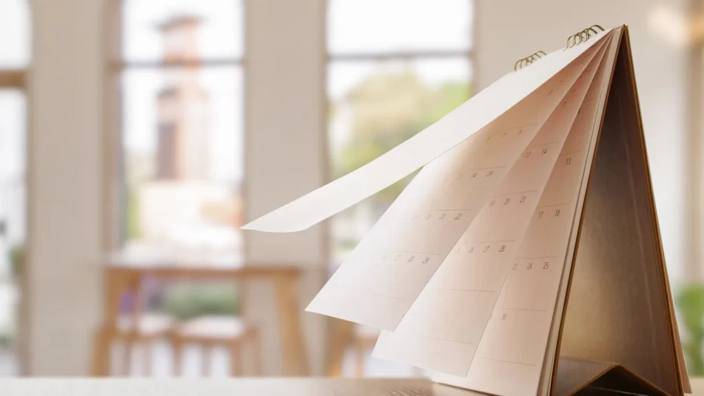 Calendrier de bureau en papier dont les pages se tournent, posé sur une table en bois devant de hautes fenêtres lumineuses