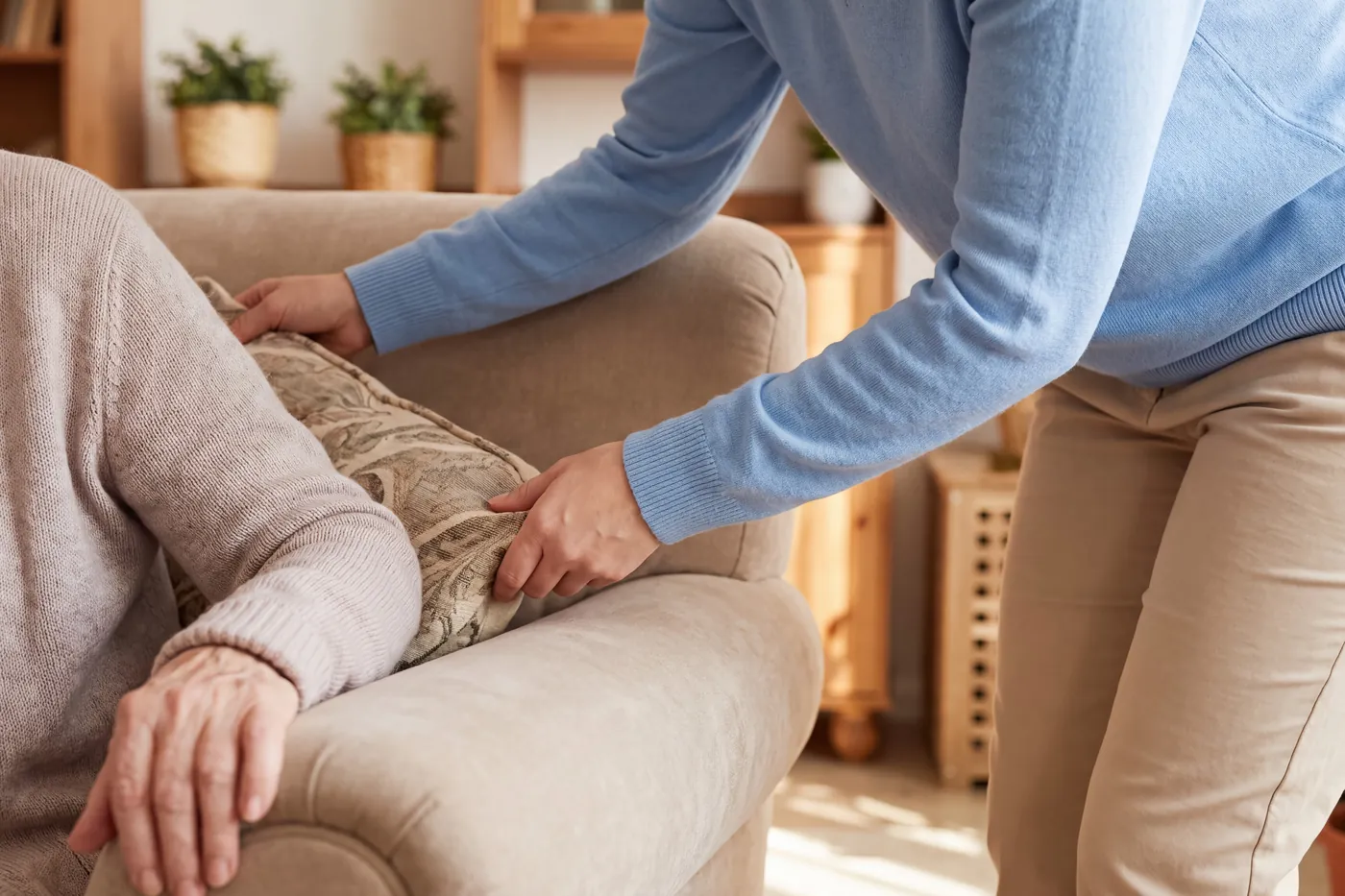 Personne ajustant un coussin pour un proche assis dans un fauteuil beige, dans un salon chaleureux, visages hors du cadre