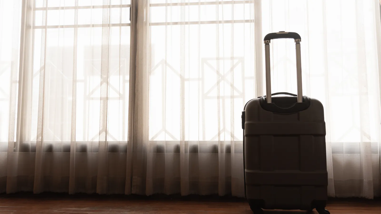 Valise cabine posée sur un parquet devant de hautes fenêtres voilées de rideaux clairs, dans une douce lumière de fin de jour
