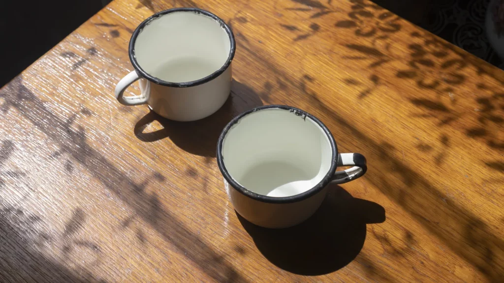 Deux tasses émaillées blanches posées sur une table en bois, baignées par la lumière chaude du soir et l'ombre de feuillages