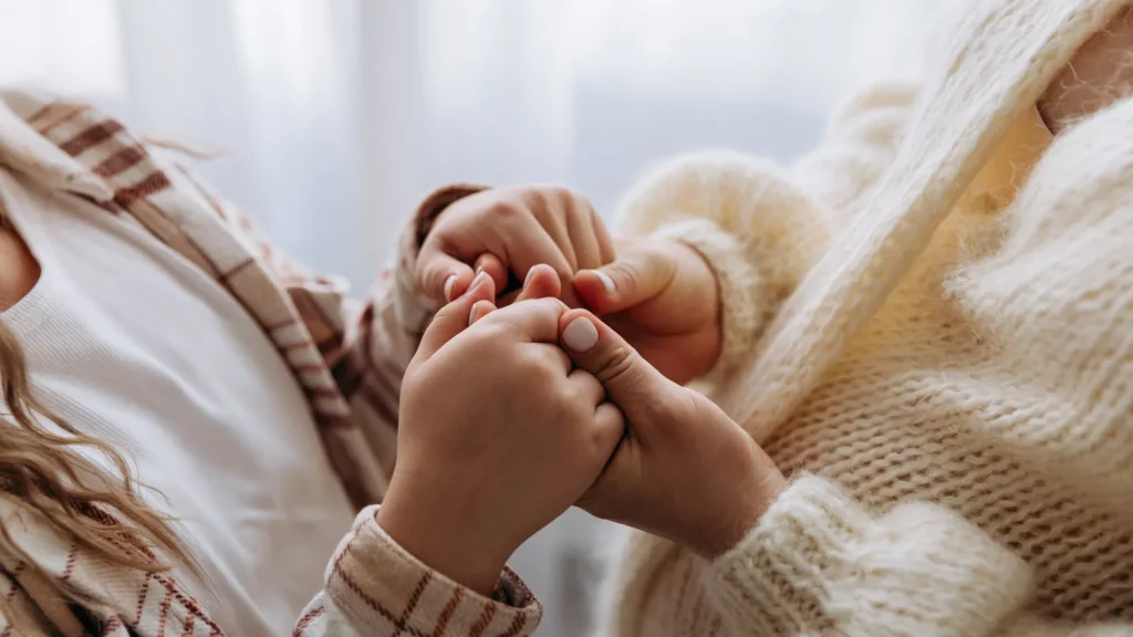 Main d'enfant serrant les doigts d'un adulte en gros plan, vêtements en maille crème, devant une fenêtre aux voilages clairs