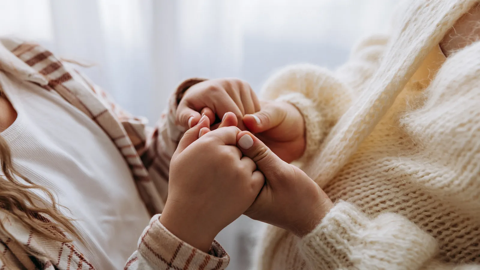 Main d'enfant serrant les doigts d'un adulte en gros plan, vêtements en maille crème, devant une fenêtre aux voilages clairs
