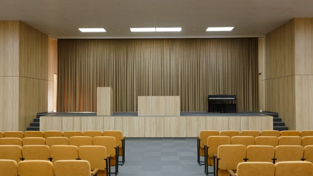Auditorium vide aux boiseries claires, rangées de sièges ocre face à une scène avec pupitre et rideau beige